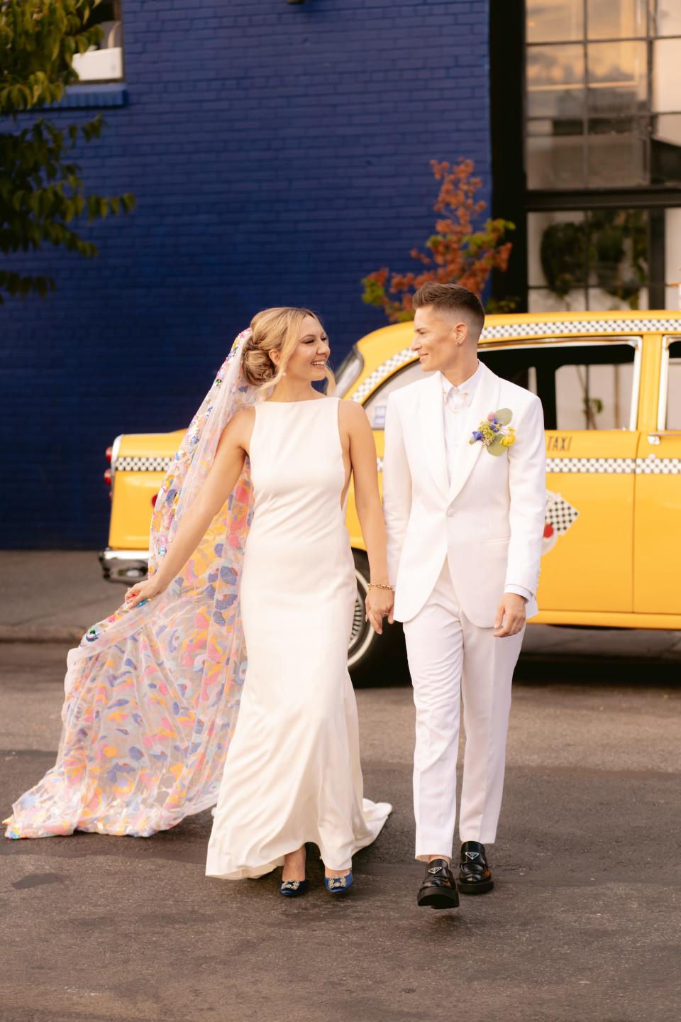 A bride in a white dress and rainbow veil and a bride in a white suit hold hands as they walk away from a yellow taxi.