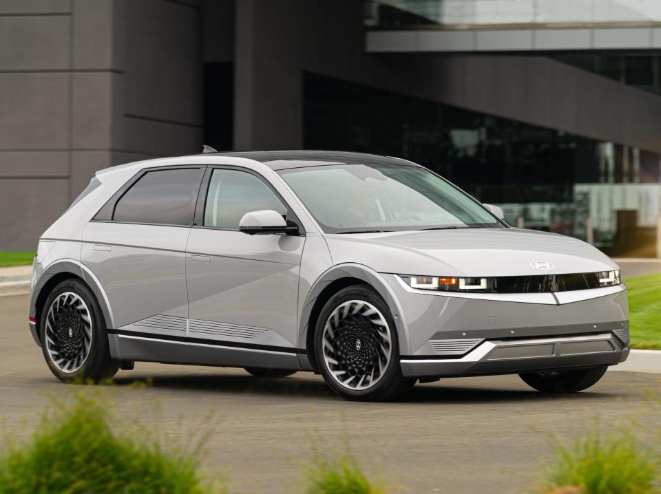 A light grey car parked on a road in front of a modern building with some surrounding greenery.