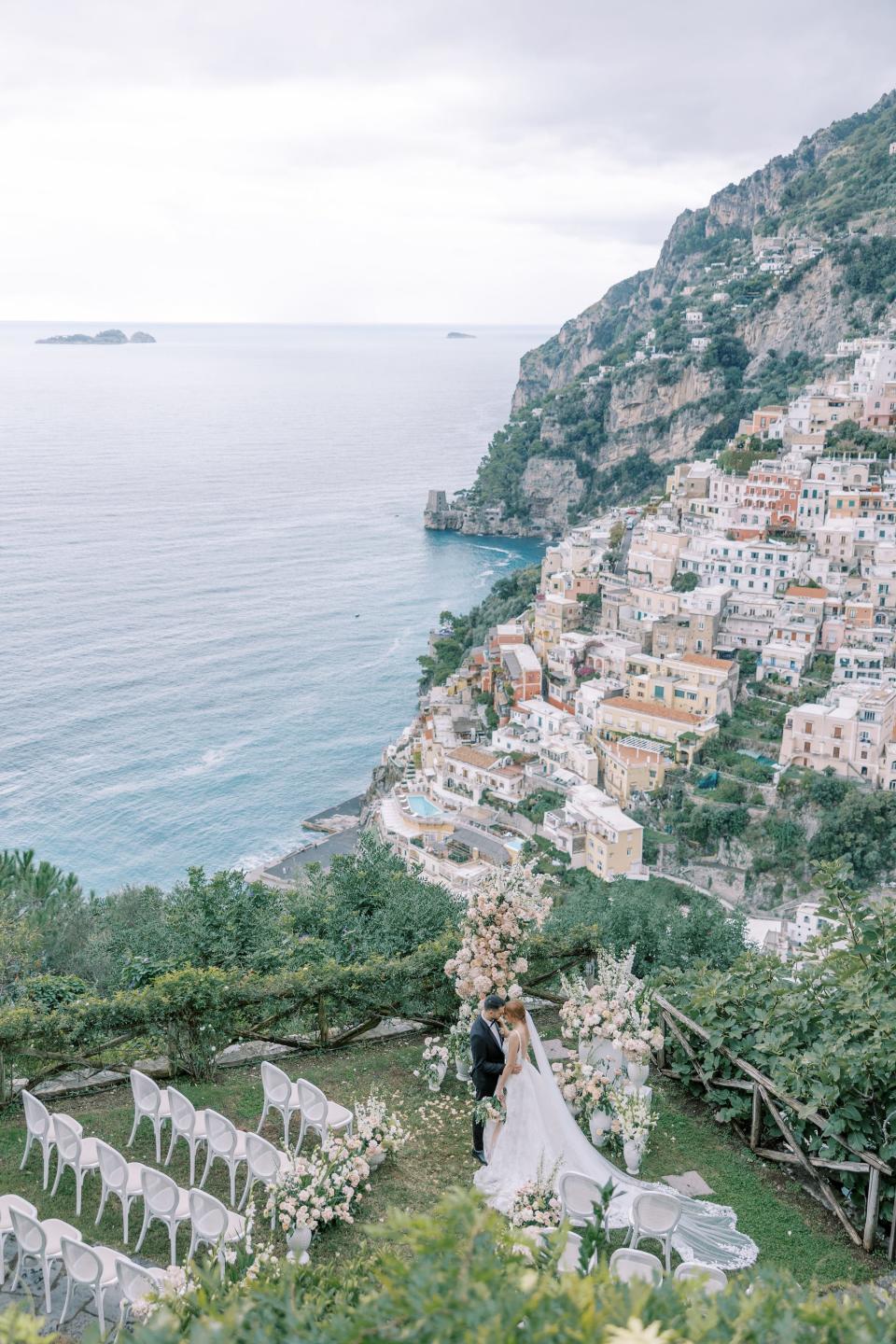 A bride and groom embrace at their wedding ceremony overlooking an ocean and town.