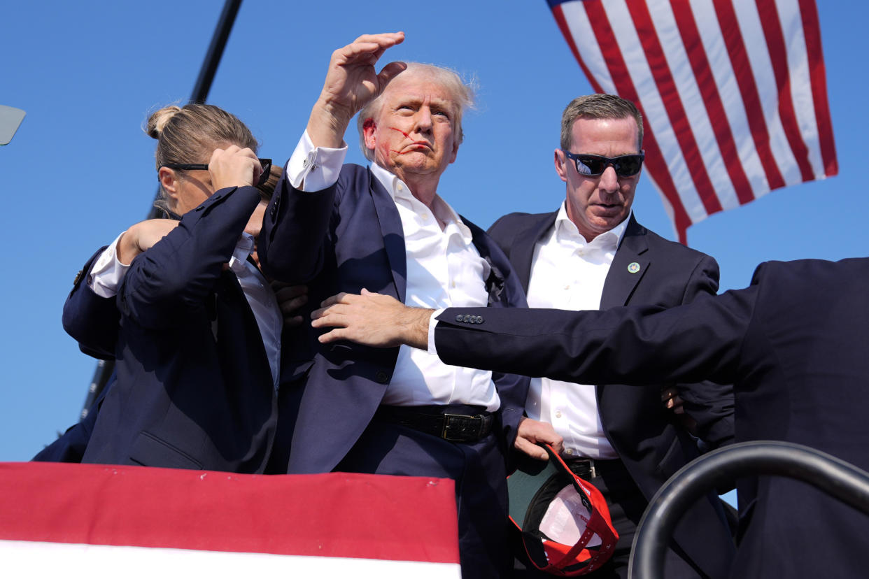  Trump, with apparent blood on his face and attempting to wave to supporters, is surrounded by Secret Service agents as he leaves the stage with their help.