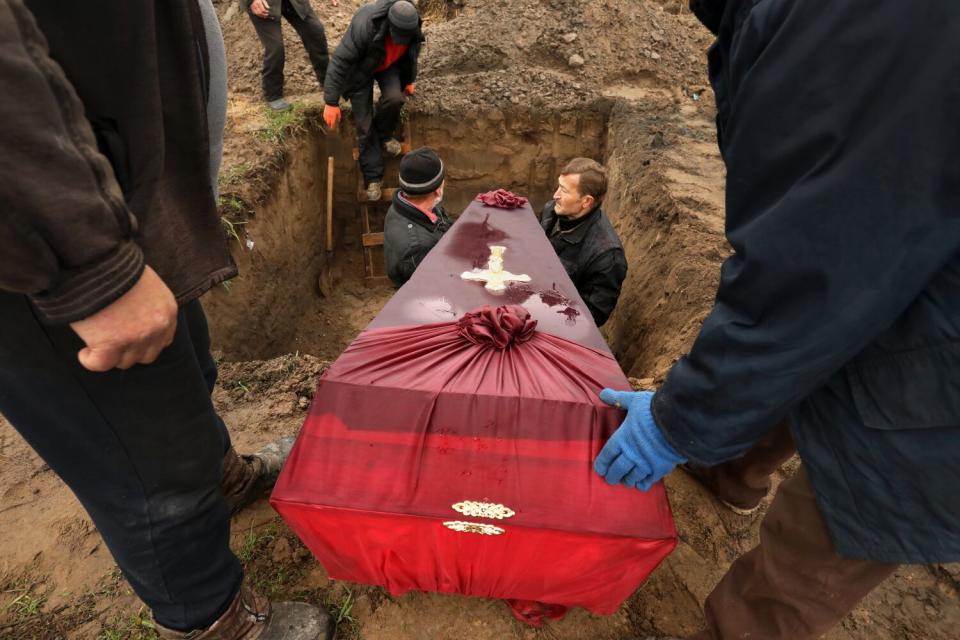 Two men stand in a large hole and hold a red casket being lowered into the grave.