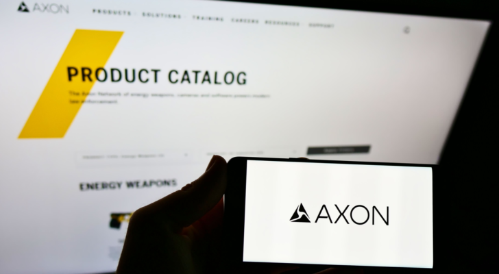 Person holding mobile phone with logo of American weapon manufacturer Axon Enterprise Inc. on screen in front of webpage. Focus on cellphone display. Unmodified photo.