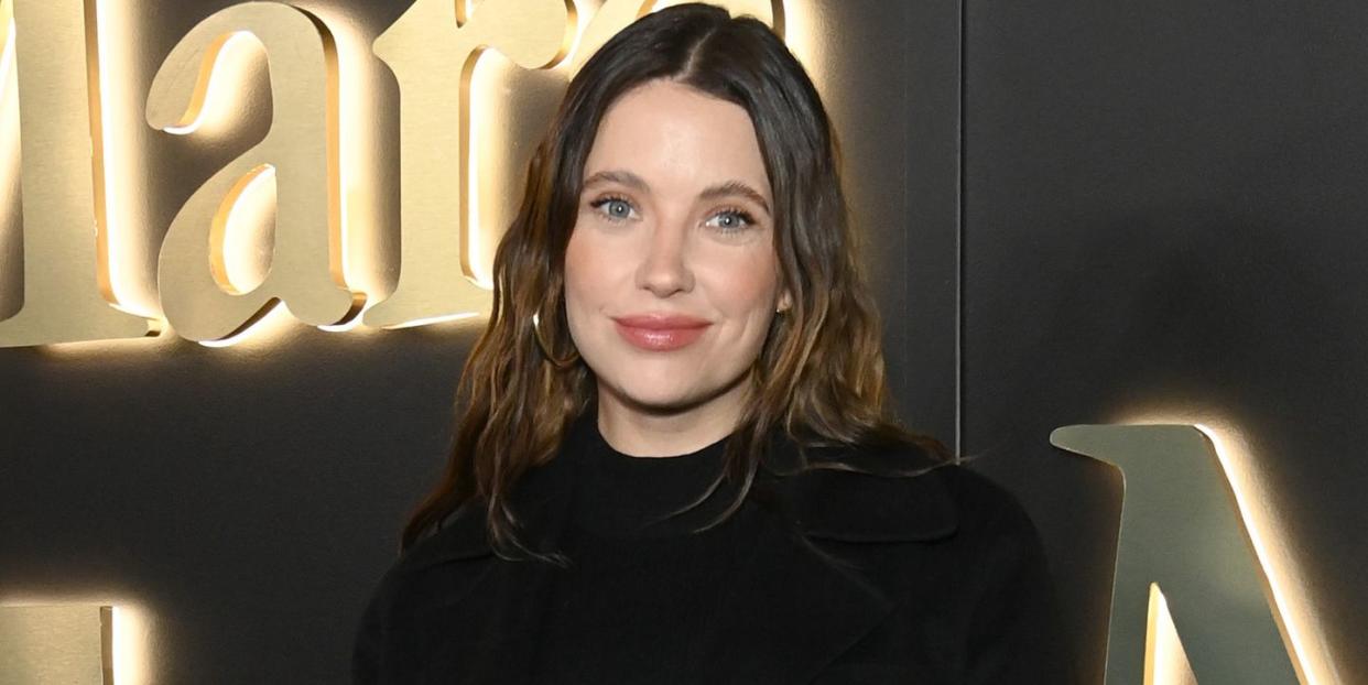 ashley benson, a woman stands smiling at the camera wearing a long black coat and with dark brown hair down in loose waves