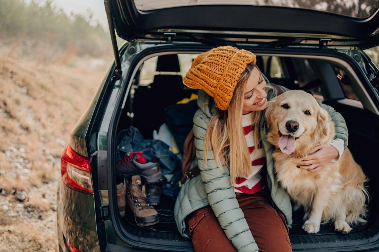 Young woman hugging her dog, both are sitting in the trunk of a SUV, during autumn, with a mound and bushes blurred in the background