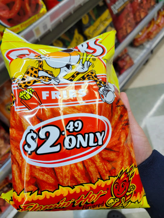 A hand holding a bag of Flamin' Hot Fries snacks, priced at just $2.49, in a grocery aisle with another bag of snacks in the background.