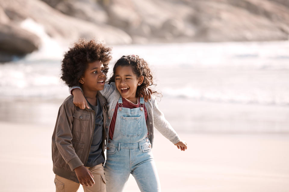 Two children walk on a beach, smiling and laughing. The one on the left wears a jacket and pants, while the child on the right wears overalls and a sweater