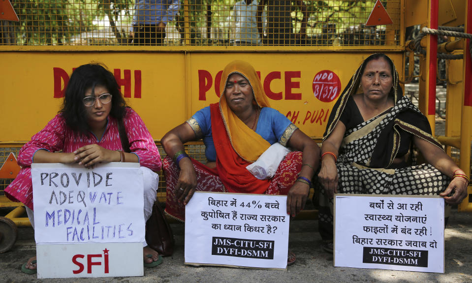 Left wing activists protesting against deaths of more than 100 children due to Encephalitis in the Indian state of Bihar, sit holding placards under a shade near a police barricade in New Delhi, India, Tuesday, June 18, 2019. (AP Photo/Altaf Qadri)