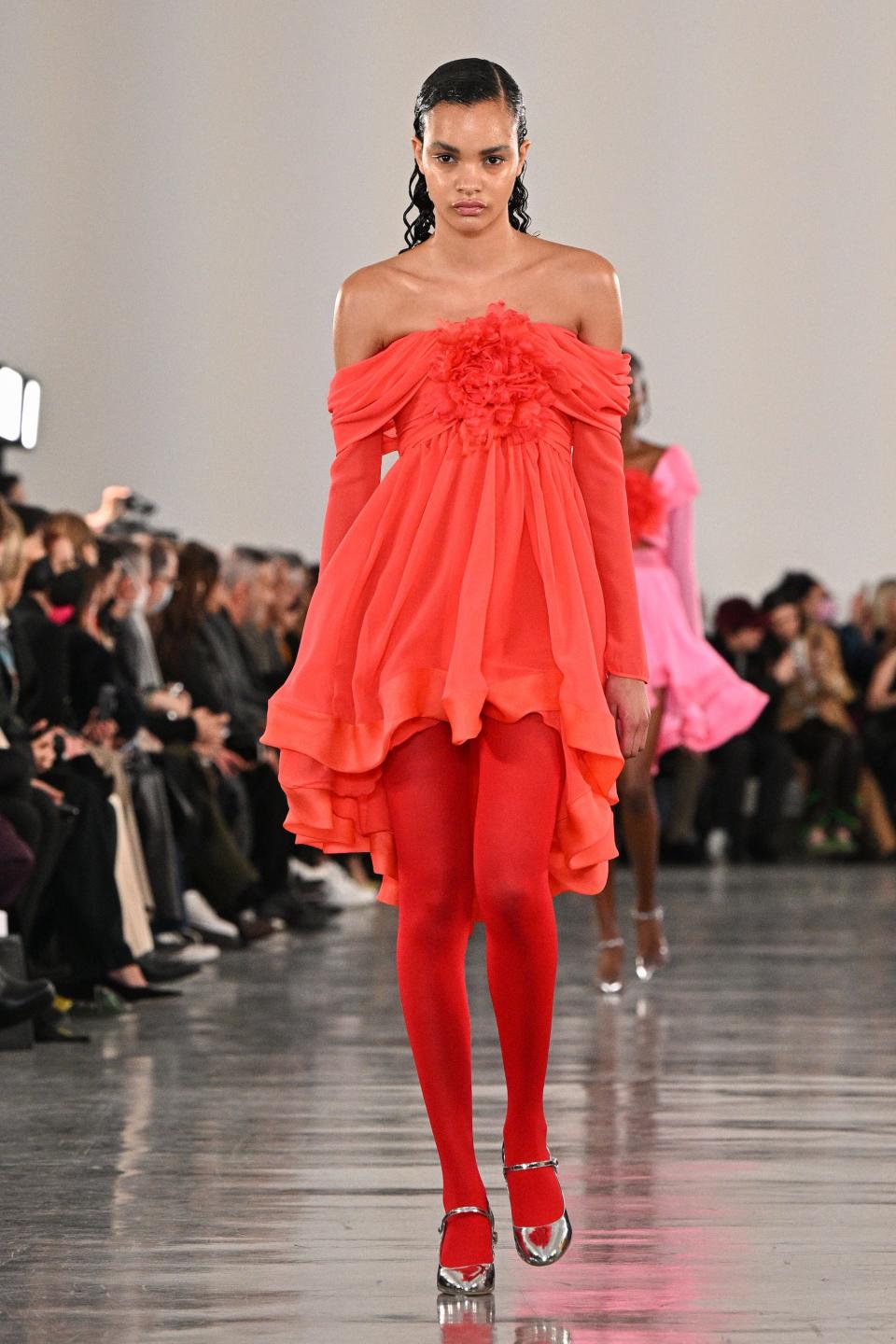 A model walks down a runway wearing an off-the-shoulder red dress with red tights and silver shoes.