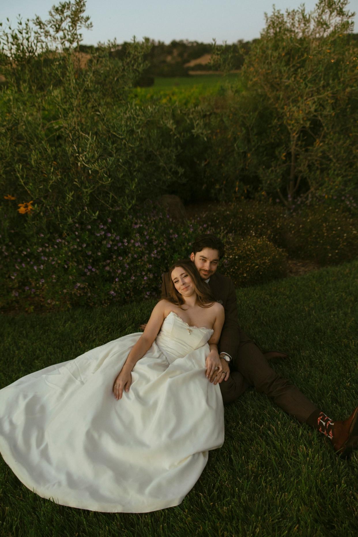 A bride and groom sit on grass together in their wedding attire.