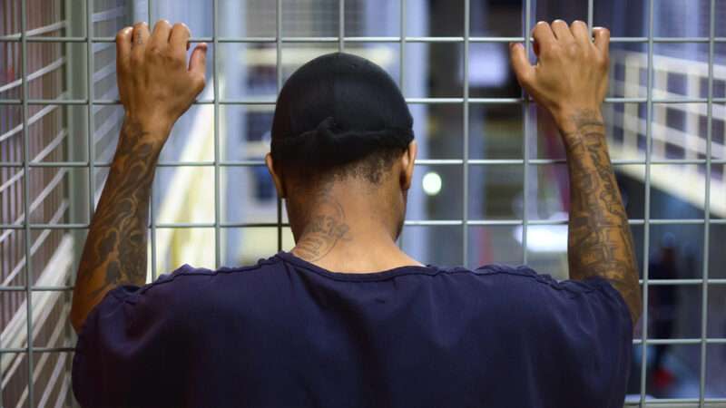 An inmate seen from behind leaning against jail cell bars