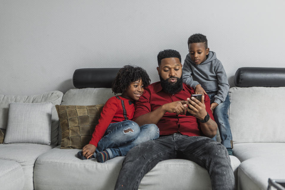 A Black father shows his two children, who sit on either side of him on a sofa, something on his phone