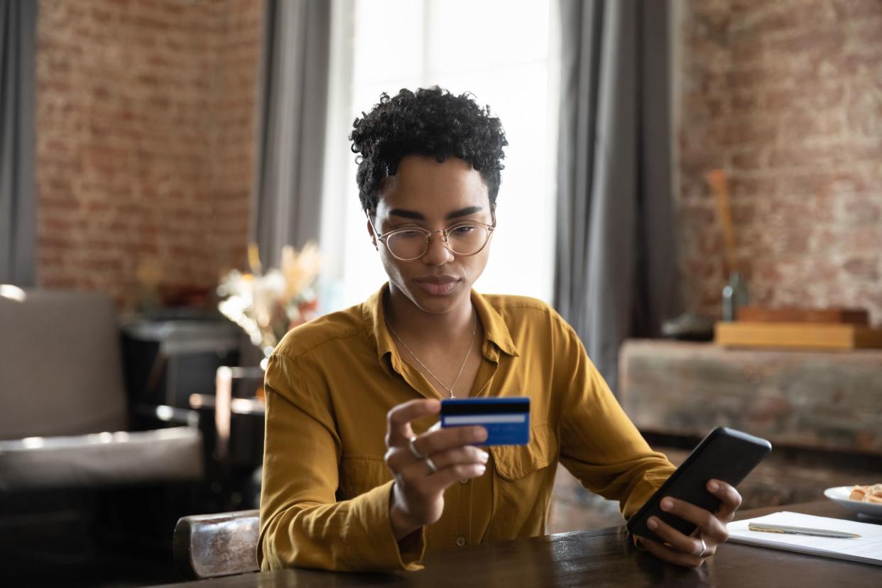 Make secure quick money transfer, e-services and retail mobile application usage, pay bills on-line concept. Serious African woman sit at desk holds smartphone and credit card, do shopping on internet