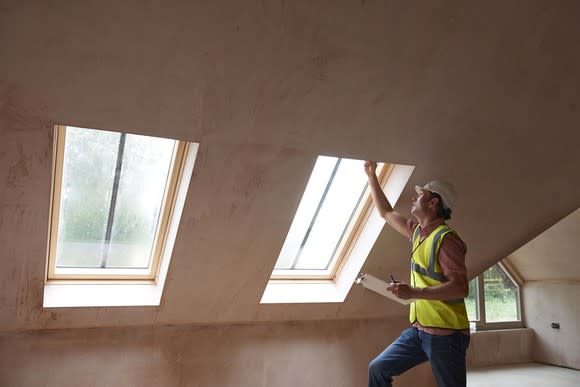 A home inspector in a yellow vest holds a clipboard as he looks at a window.