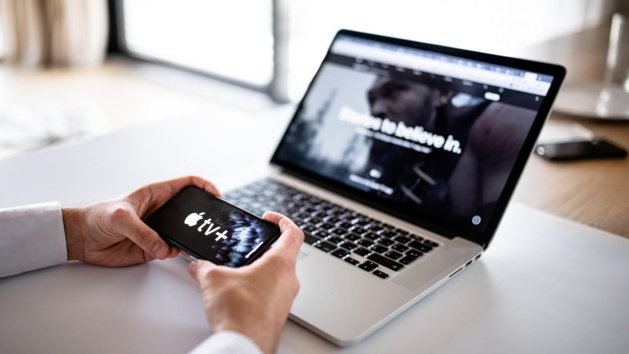  Hands hold smartphone with Apple TV Plus logo, next to a MacBook with Apple TV Plus also on it. 