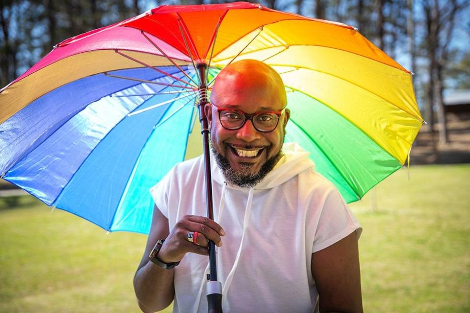 A smiling bisexual black man living with HIV holding a colorful rainbow umbrella on a sunny day