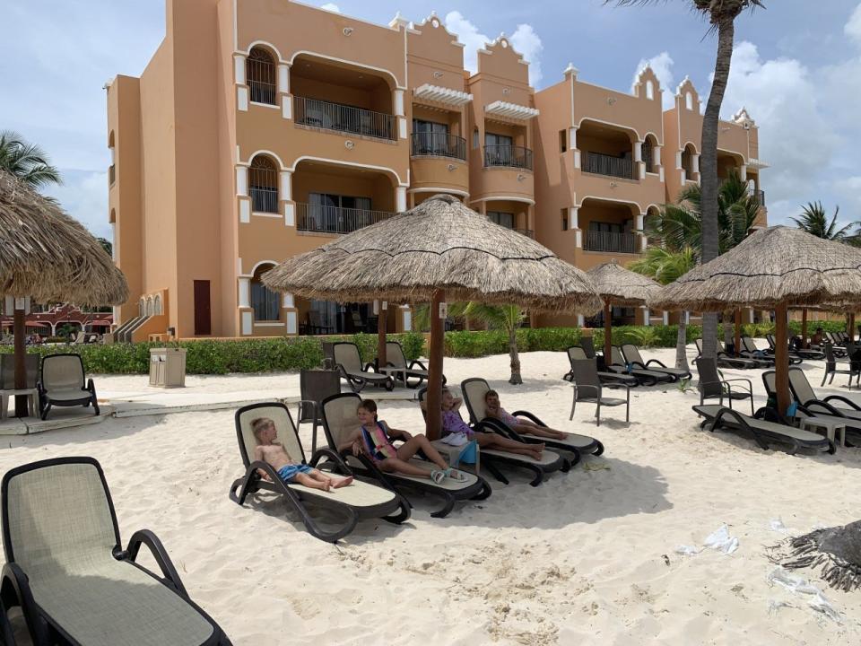 Four children sitting on beach chairs under a palapa umbrella at the beach in front of a hotel.