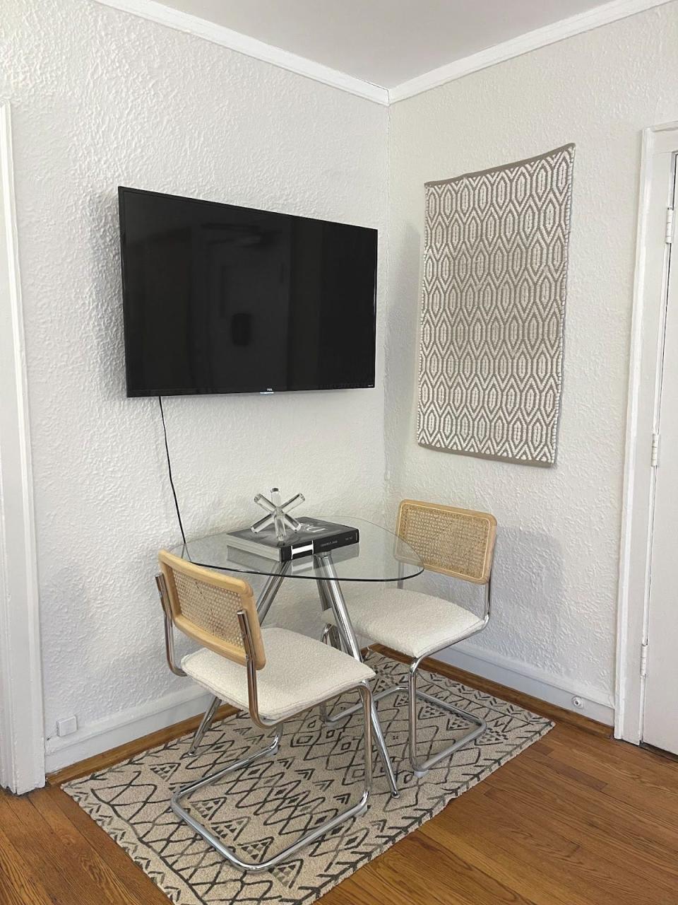 A tv on a light wall above a small dining set with a glass table and rug beneath it, plus a gray rug hanging on the wall