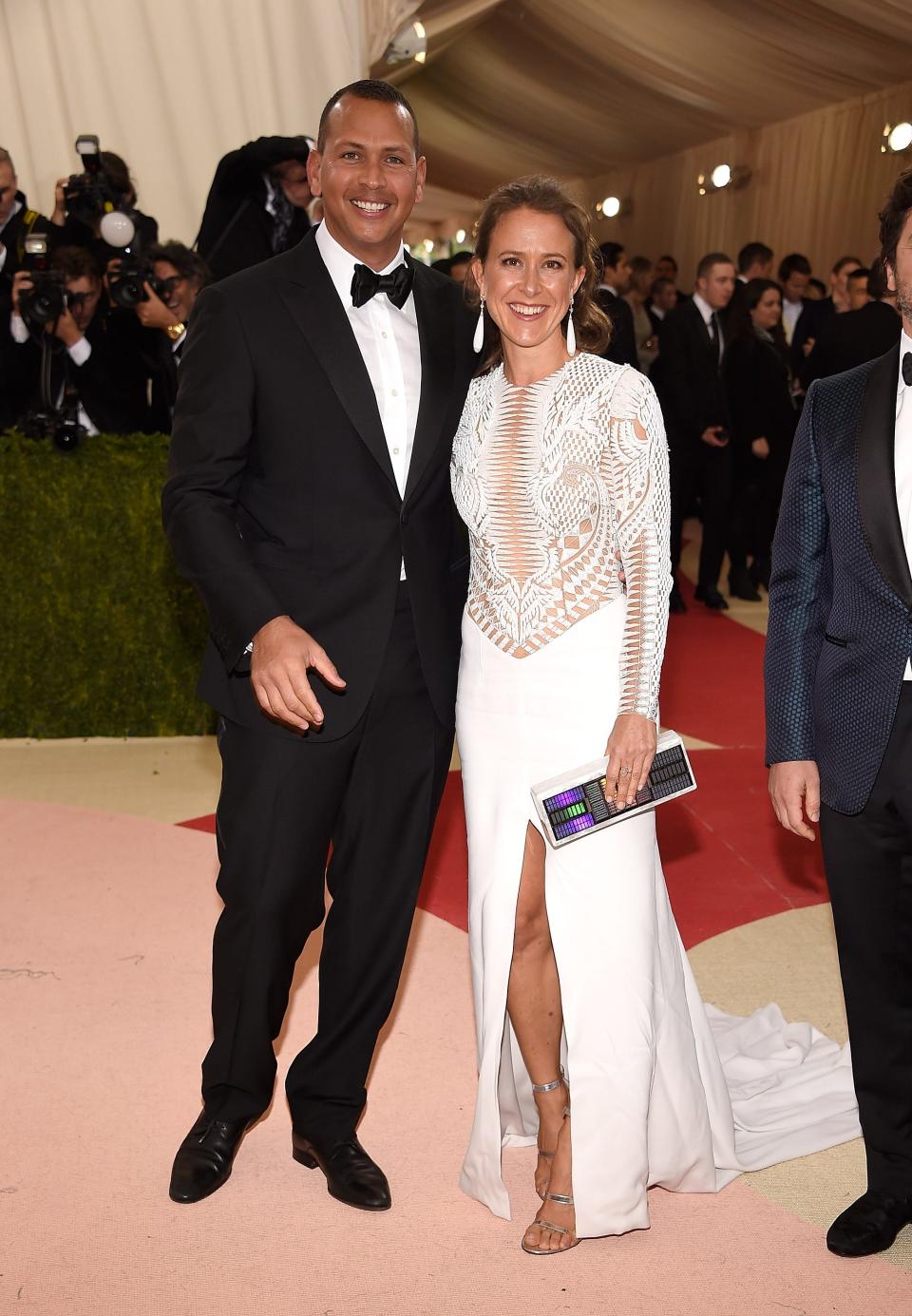 Alex Rodriguez stands in a suit and Anne Wojcicki stands in a white dress on a red carpet.