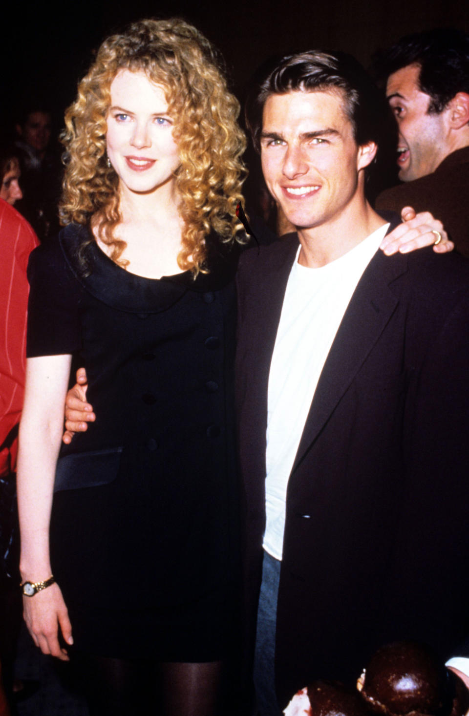 Nicole Kidman and Tom Cruise at an event, smiling for the camera. Nicole is wearing a black dress, and Tom is in a white shirt with a black blazer