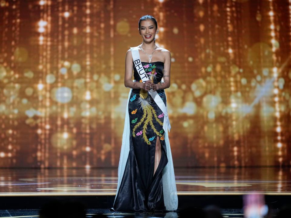 Miss Bhutan 2022, Tashi Choden, stands in a patterned black evening dress and sash in front of a glittering gold backdrop on stage in during the preliminary round of the 71st Miss Universe Beauty Pageant in New Orleans, Wednesday, Jan. 11, 2023.