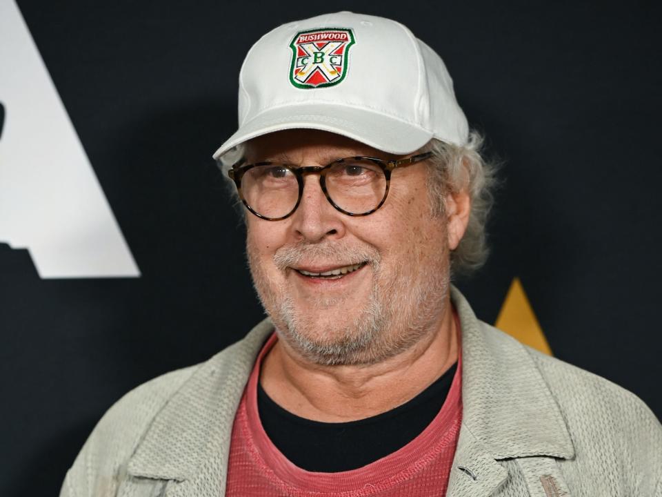 Chevy Chase at an event in 2019. He wears a red shirt under a gray jacket and a baseball hat.