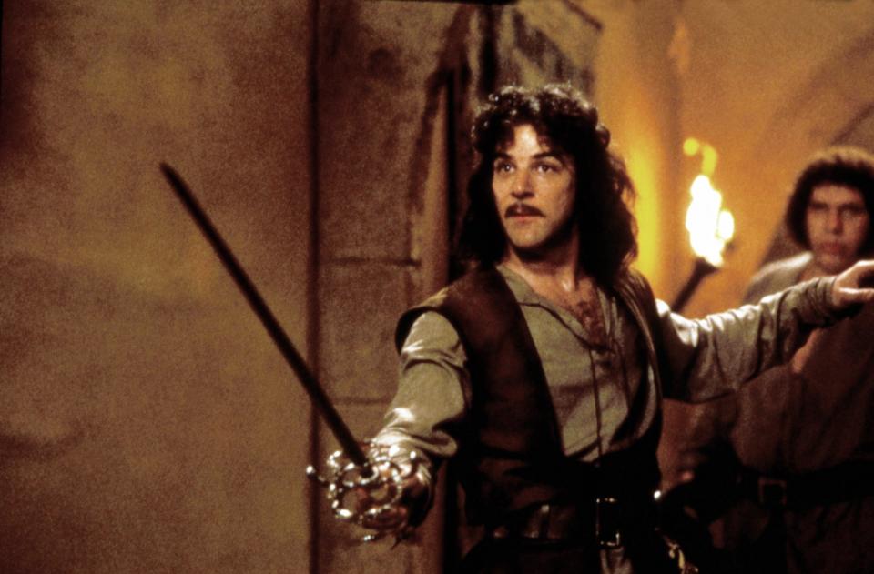 Inigo Montoya, a character from 'The Princess Bride,' is holding a sword and looking determined, with a stone wall and a torch in the background