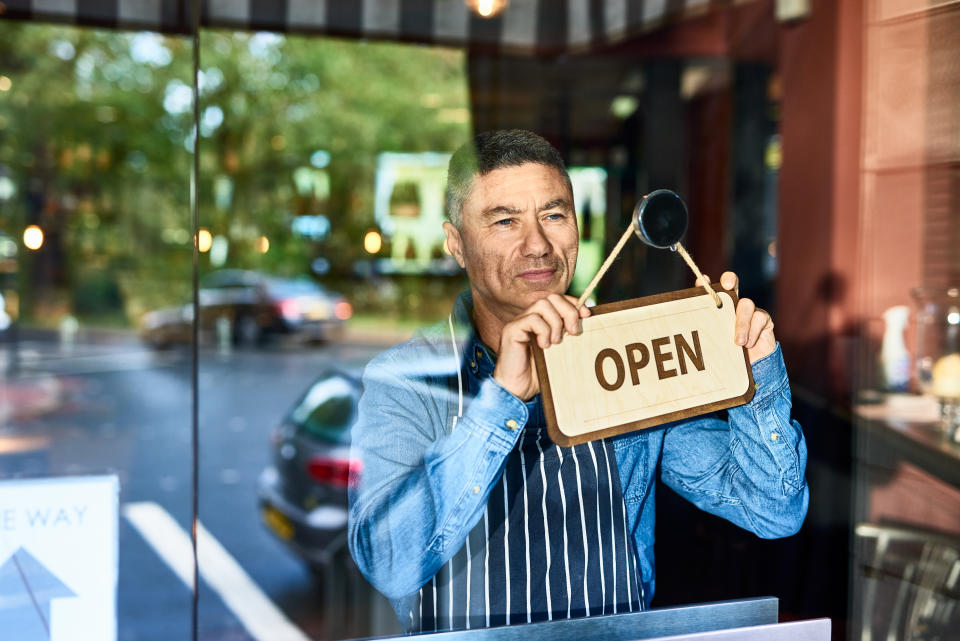 Mature man in his 40s holding open sign on door of business, looking through window, optimism, aspiration, resilience