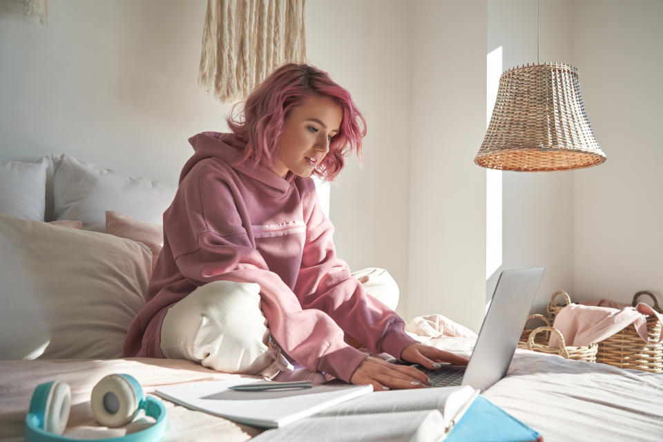 Hipster teen girl school student with pink hair wearing hoodie using laptop computer sitting in bed distance elearning online learning course for exam search remote online classes in bedroom at home.