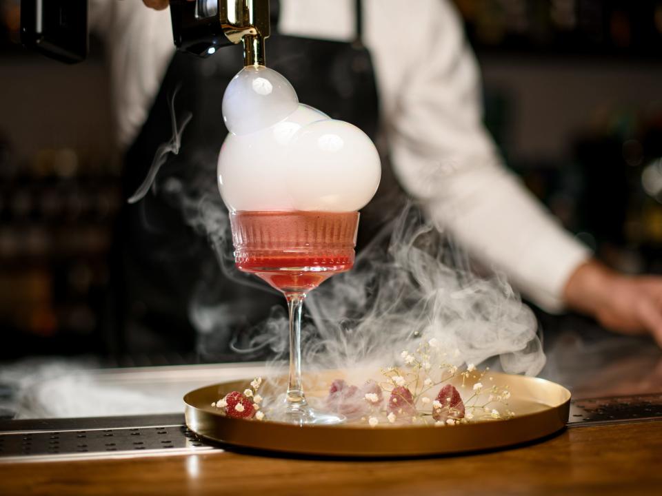 Barkeeper, der mit einer Rauchpistole einen rosa Cocktail in einer Bar präsentiert.