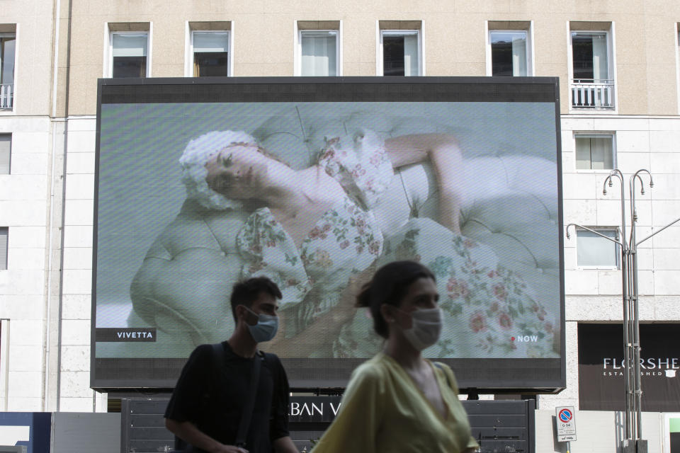 Pedestrians pass by a screen showing a Vivetta model during the Milan Digital Fashion Week, in Milan, Italy, Tuesday, July 14, 2020. Forty fashion houses are presenting previews of menswear looks for next spring and summer and pre-collections for women in digital formats, due to concerns generated by the COVID-19. (AP Photo/Luca Bruno)