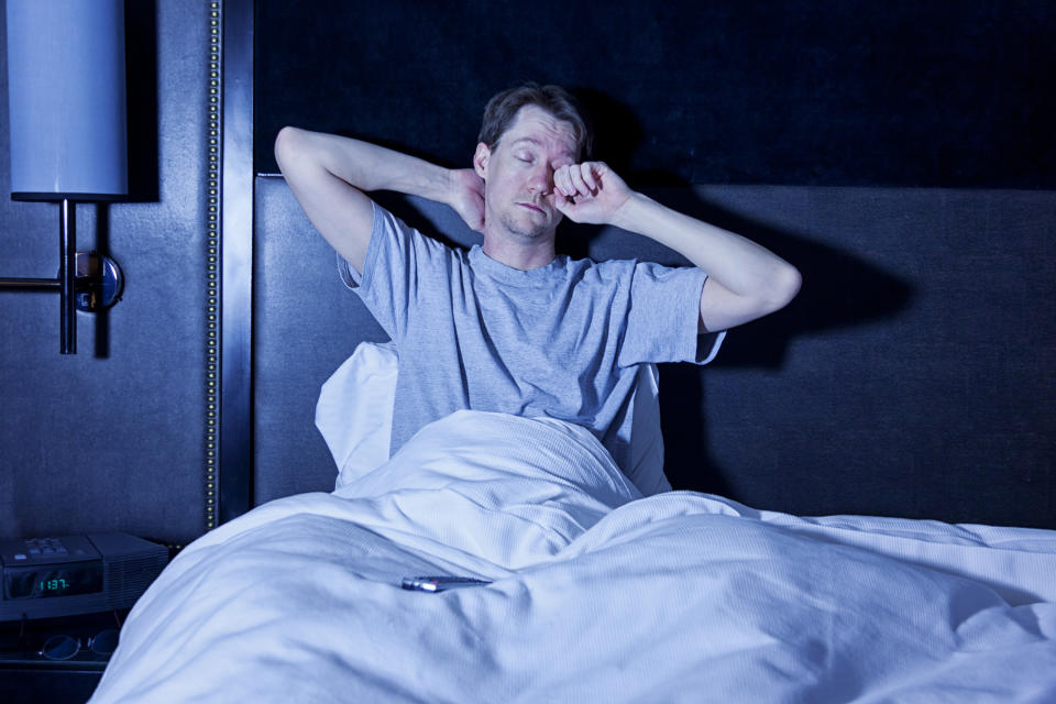 Photo of a man sitting in a hotel room bed bed, rubbing his tired eyes as he watches late night television with the remote control in his lap.