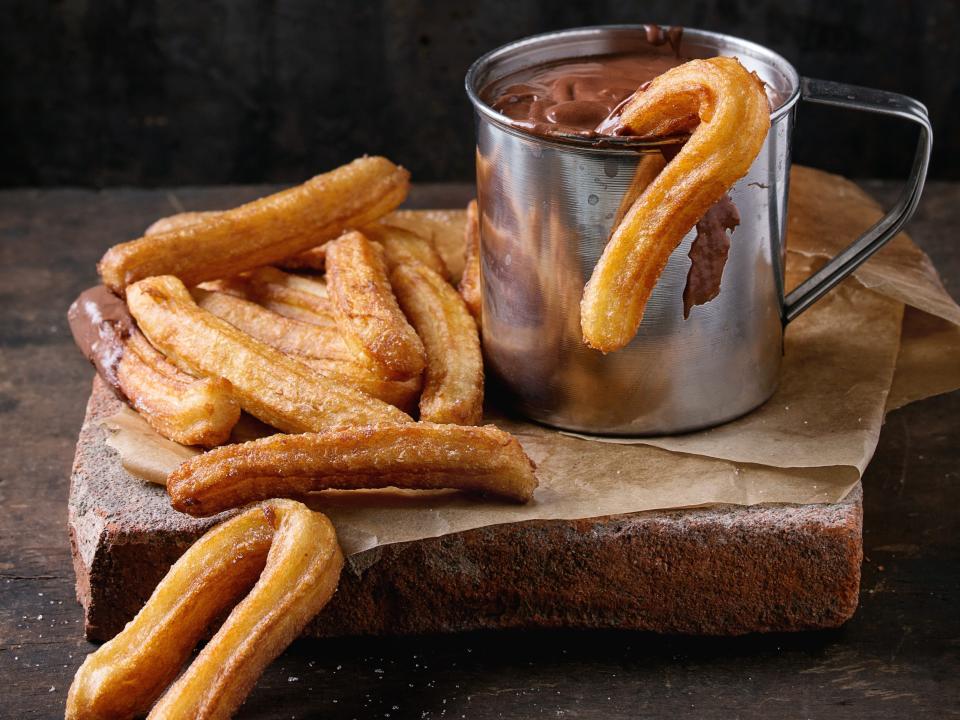 Traditional spanish treat churros with chocolate sauce in metal mug, served on baking paper over dark wooden background.