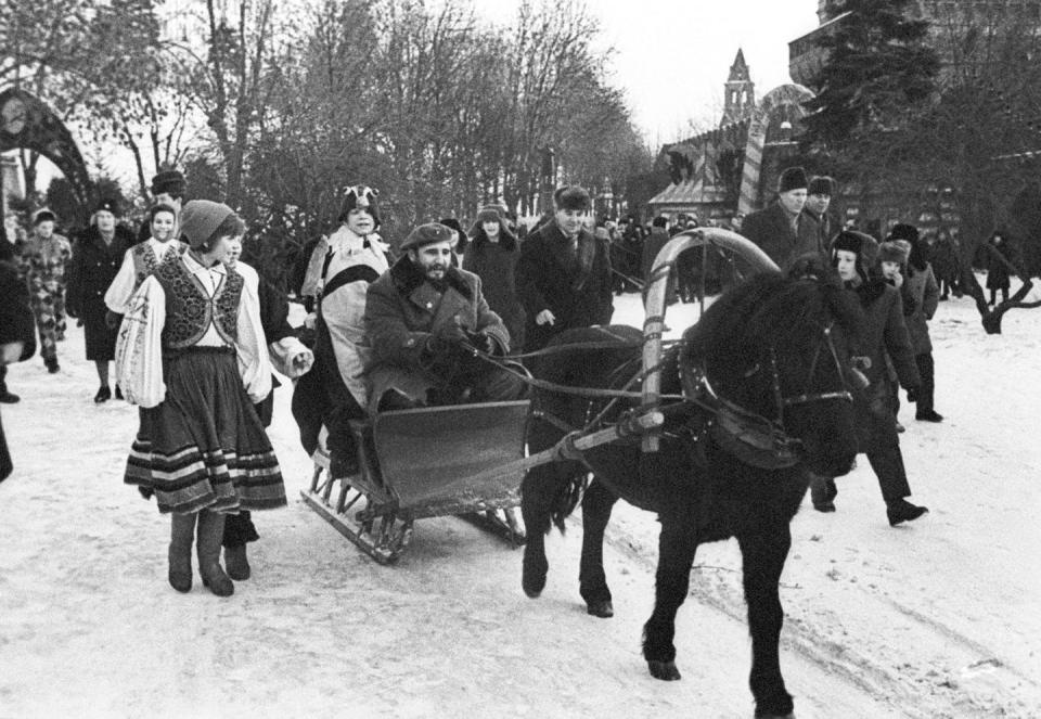 Black-and-white photo of Castro, in his traditional revolutionary beret, driving a horse-drawn sleigh in the snow, surrounded by Russians in traditional dress