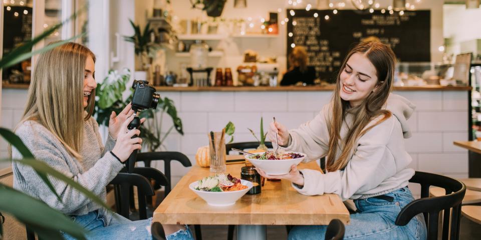 Two teenagers or Gen Zers in a cafe eating food. One is taking a photo of the other.