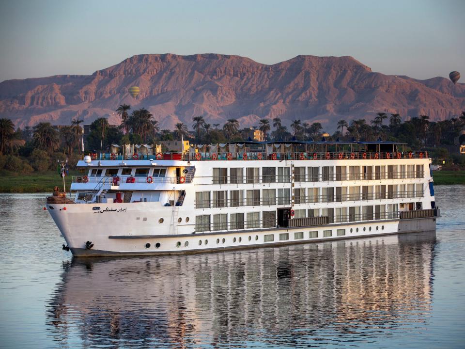 A cruise ship with several rooms with a mountain range in the background.