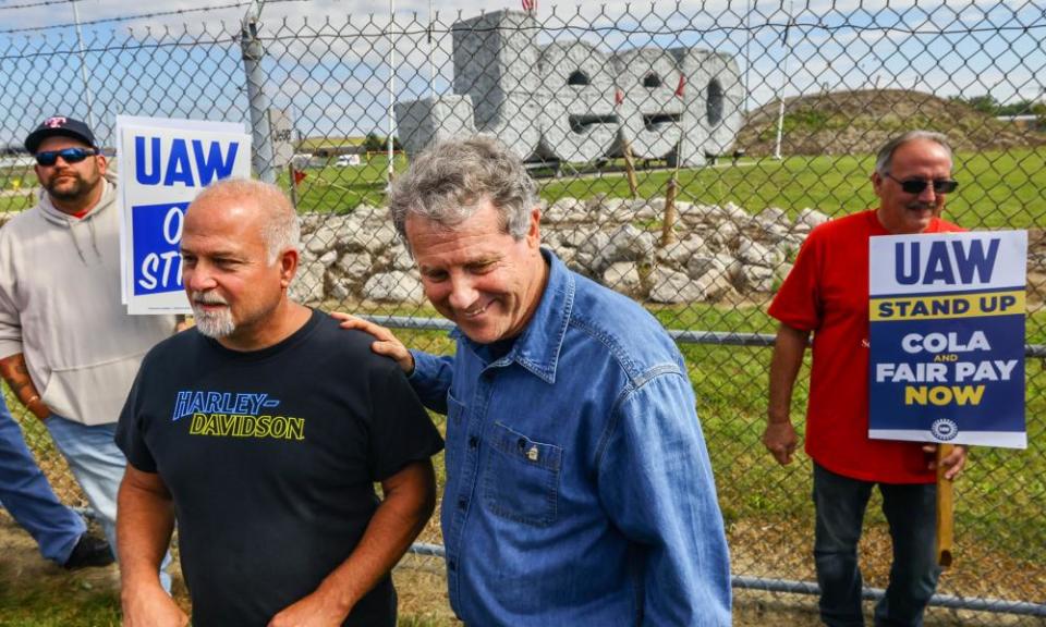 A middle-aged white man with curly gray hair wearing a blue denim shirt smiles as he has his hand on the back of a balding man with a T-shirt, among other men holding UAW protest signs, beyond a chain-link fence and a massive structure that spells out the Jeep logo.