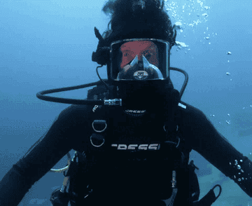 A person scuba diving underwater with an expression of surprise, wearing full scuba gear including a mask and a Cressi wetsuit