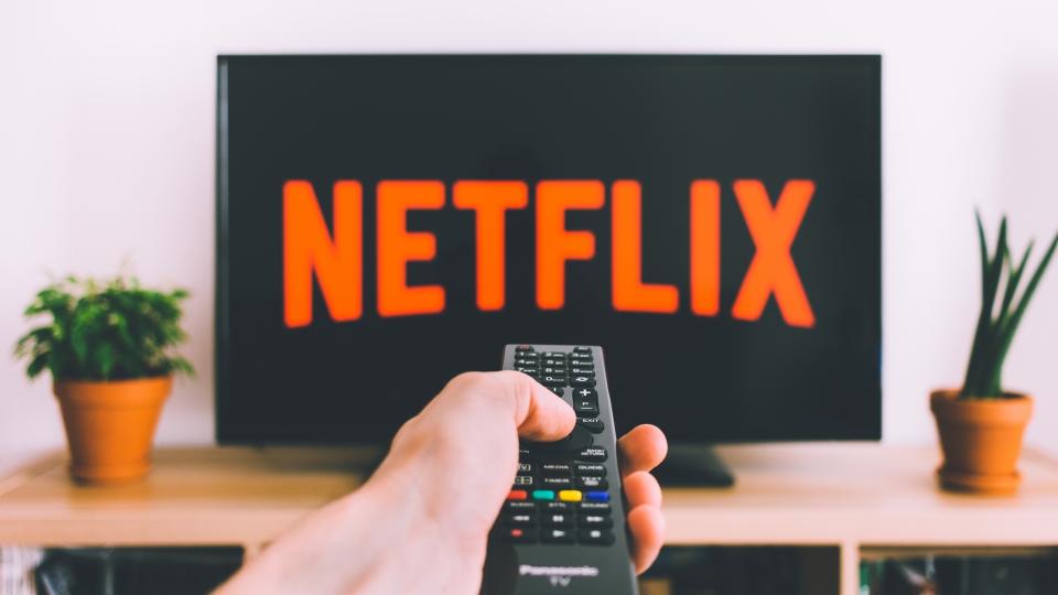  A TV with the Netflix logo sits behind a hand holding a remote. 