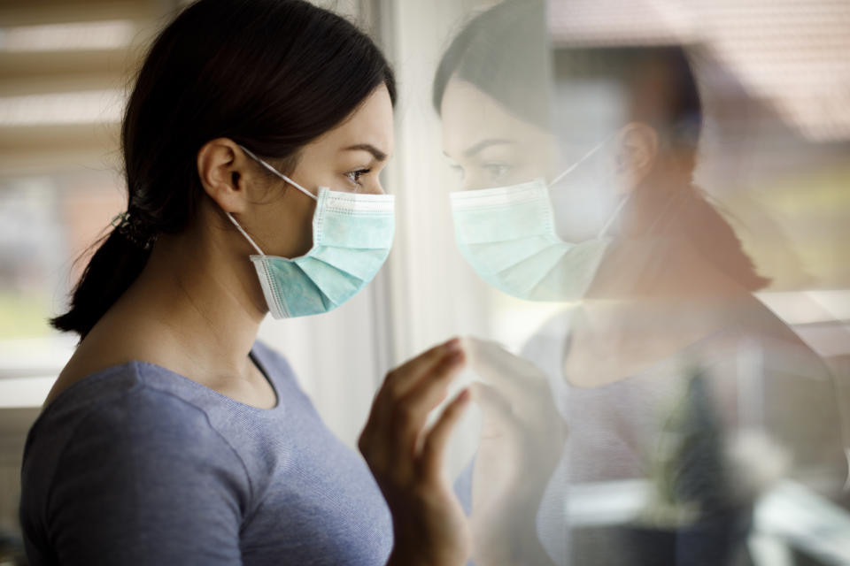 Portrait of sad young woman with face protective mask looking through the window at home