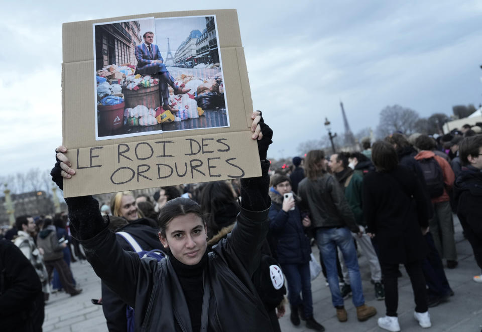 A woman holds a placard depicting French President Emmanuel Macron sitting on garbage cans that reads, "king of trash" during a protest in Paris, Friday, March 17, 2023. Protests against French President Emmanuel Macron's decision to force a bill raising the retirement age from 62 to 64 through parliament without a vote disrupted traffic, garbage collection and university campuses in Paris as opponents of the change maintained their resolve to get the government to back down. (AP Photo/Lewis Joly)
