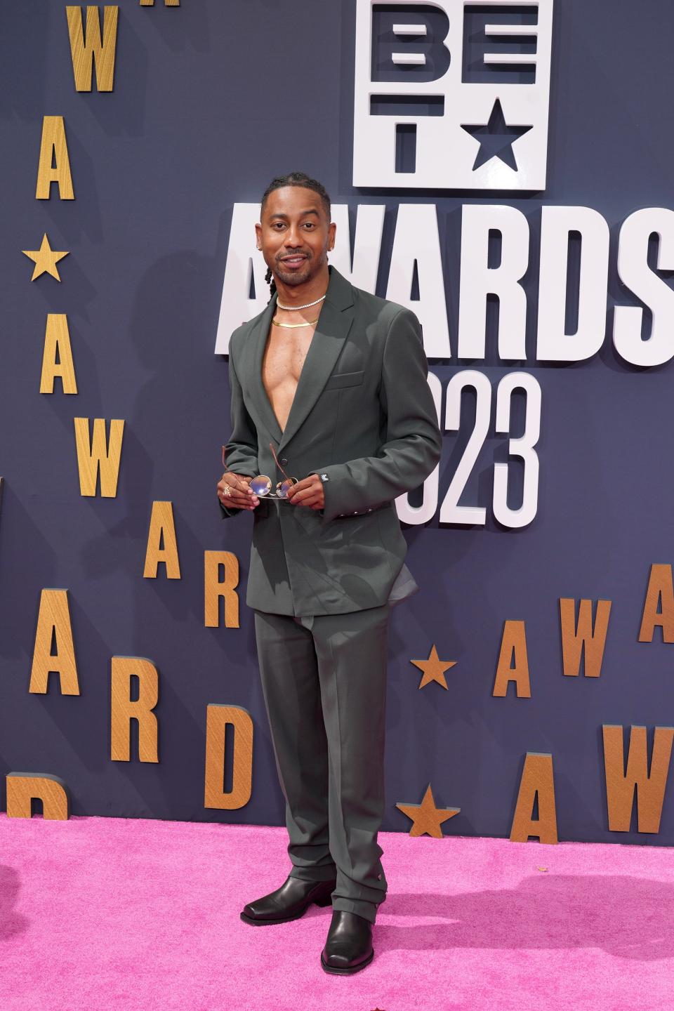 Brandon T. Jackson poses on the 2023 BET Awards red carpet in an olive suit without a shirt underneath, two necklaces, and a pair of black shoes.