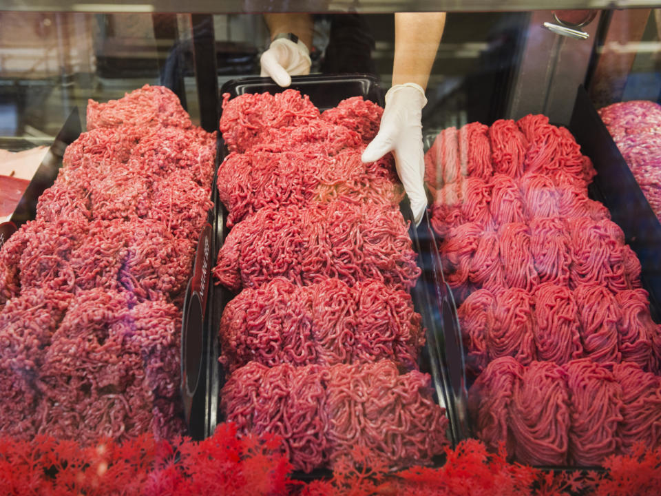 A person wearing gloves arranges trays of ground meat in a butcher's display case