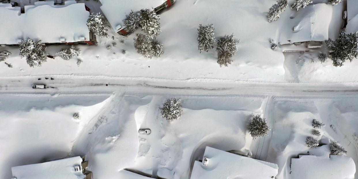 Aerial view shows a vehicle and people navigate a snowy roadway lined with snowbanks piled up from new and past storms in the Sierra Nevada mountains.