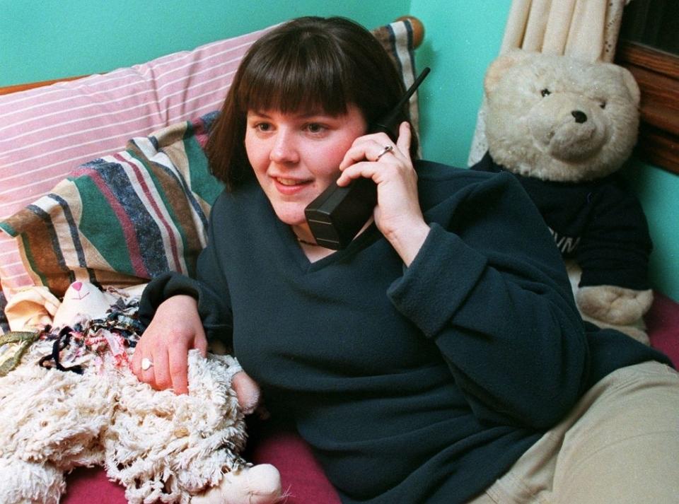 A person is sitting on a bed, holding a cordless phone, and surrounded by stuffed animals and a pillow