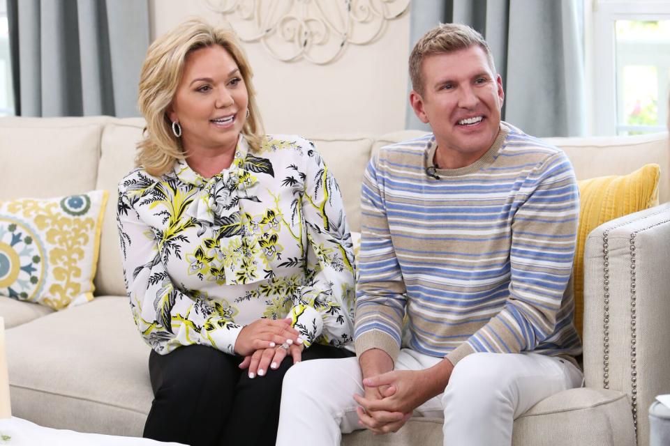 Julie Chrisley, left, in a floral shirt and black pants sitting next to Todd Chrisley in a striped shirt and white pants