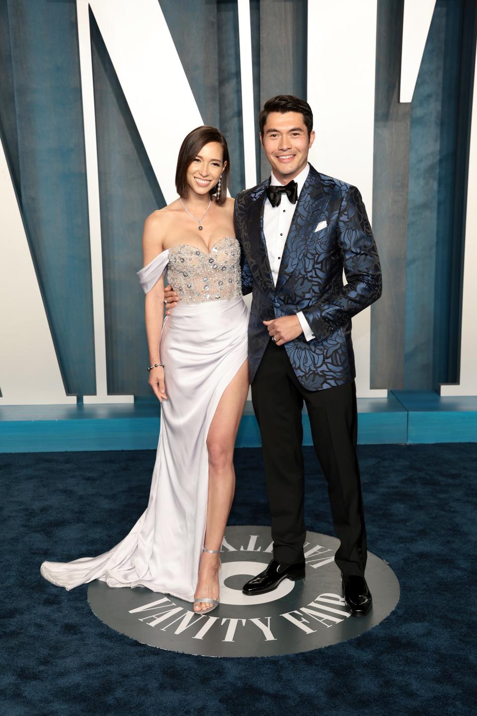 Liv in a pale blue-silver gown. The bodice is sheer showing large sequence and boning. The off the shoulder mini skeeves and thigh-high slit skirt are satin. Henry is in a dark blue shiny brocade blazer with a black bowtie and pants.