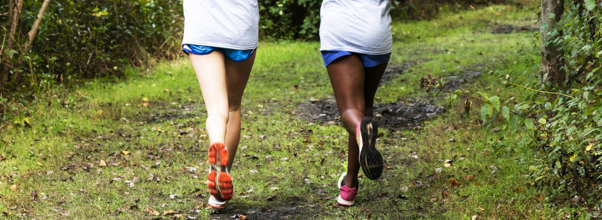 Two high school cross country runners are running side by side on a green path in the woods during practice.