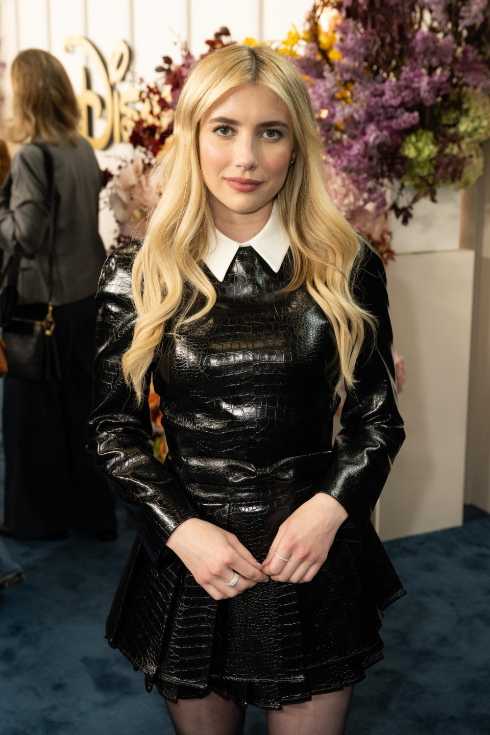 Emma Roberts wearing a black textured dress with a white collar at an event. She stands in front of a backdrop with flowers. Other attendees are in the background
