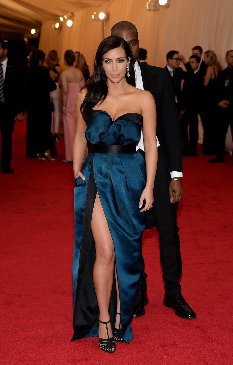 Kim Kardashian on the Met Gala red carpet wearing a blue gown with a thigh-high slit. Her then-husband Kanye West stands directly behind her.