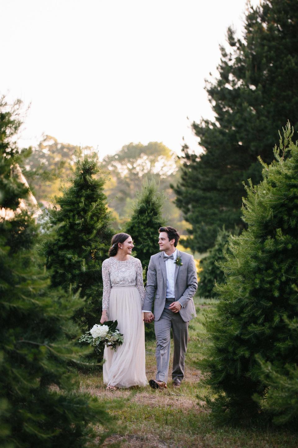 A bride and groom smile at each other as they walk through rows of Christmas trees.